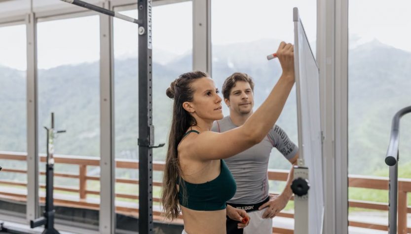 Female fitness coach writes on whiteboard as a male athlete watches in a gym.